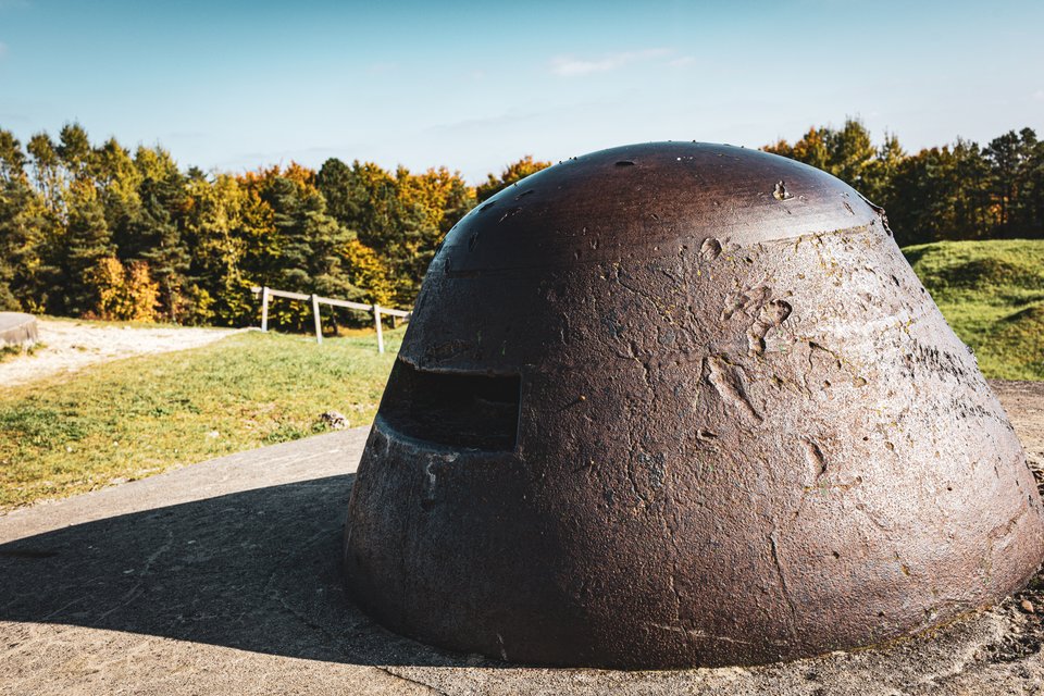 A large rusted steel military bunker dome with an observation slit, set in a concrete platform on a grassy field with trees in the background under a blue sky.
