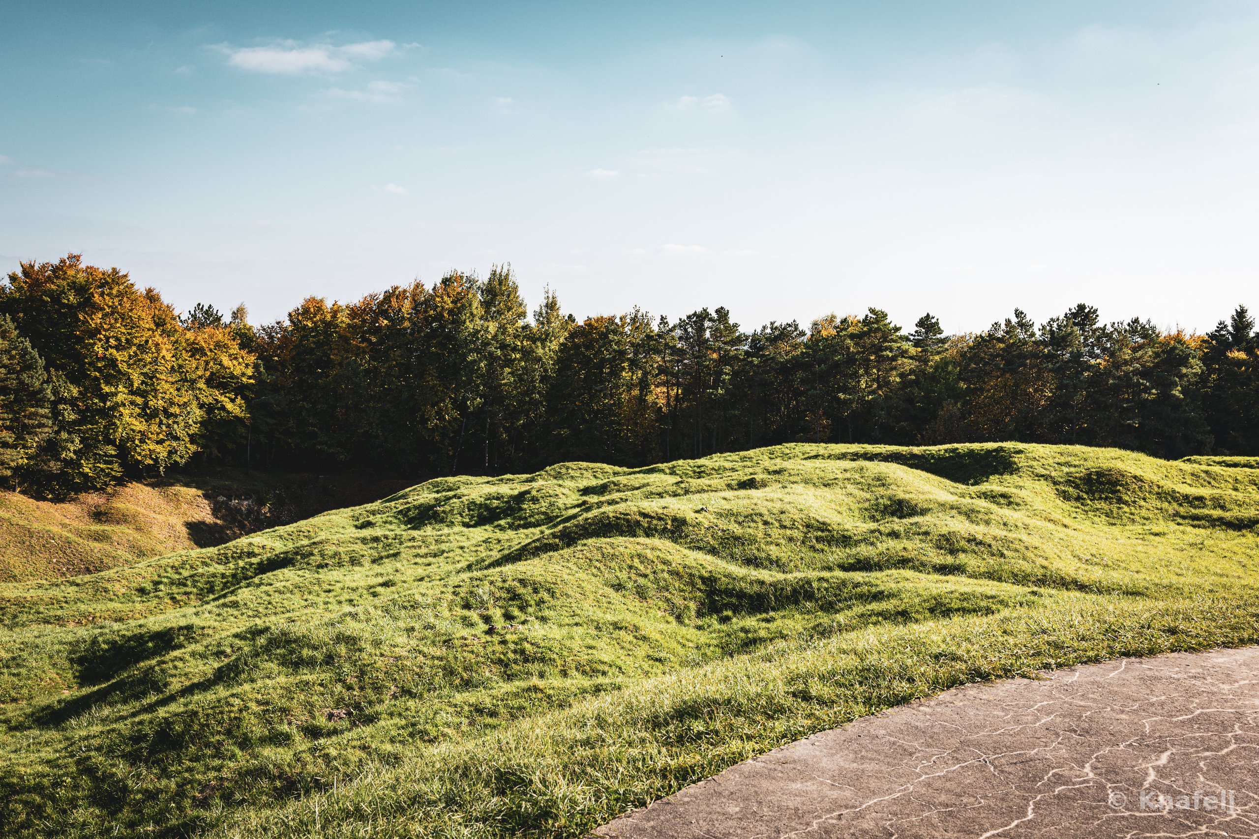 Undulating green hills with shell-crater-like depressions in front of a forest line under a pale blue sky, with a paved concrete area in the lower right corner.