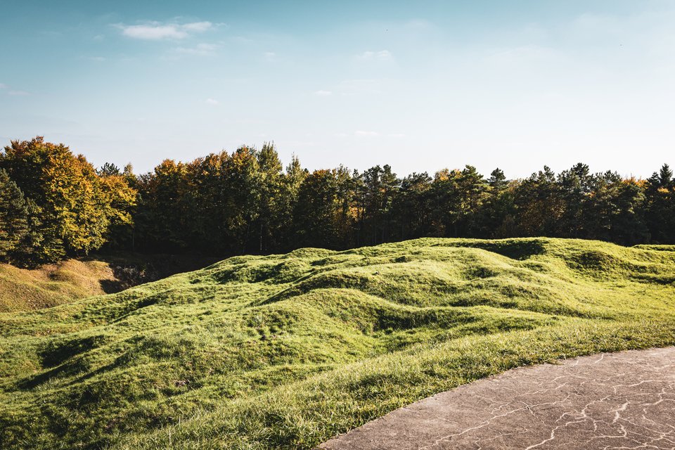 Undulating green hills with shell-crater-like depressions in front of a forest line under a pale blue sky, with a paved concrete area in the lower right corner.