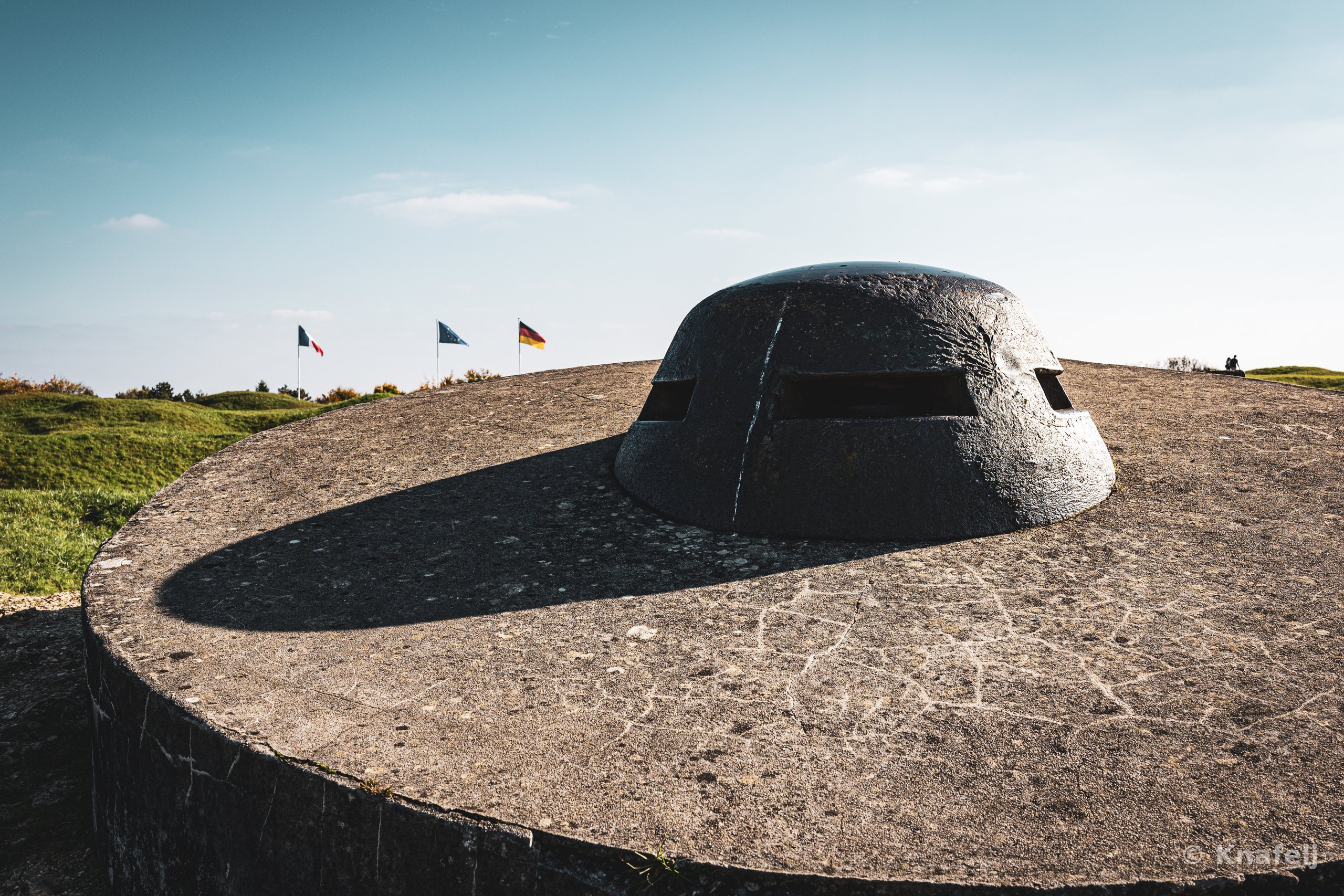 A massive rounded concrete bunker with viewing slits sits atop a cracked concrete fortification, casting a curved shadow, with three flags and green hills visible in the background beneath a pale sky.