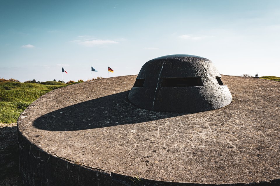 A massive rounded concrete bunker with viewing slits sits atop a cracked concrete fortification, casting a curved shadow, with three flags and green hills visible in the background beneath a pale sky.