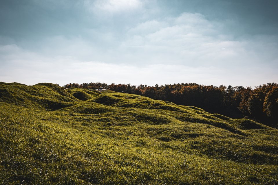 A landscape of grassy rolling hills and earthen mounds leading to a row of trees on the horizon under a partly cloudy sky.