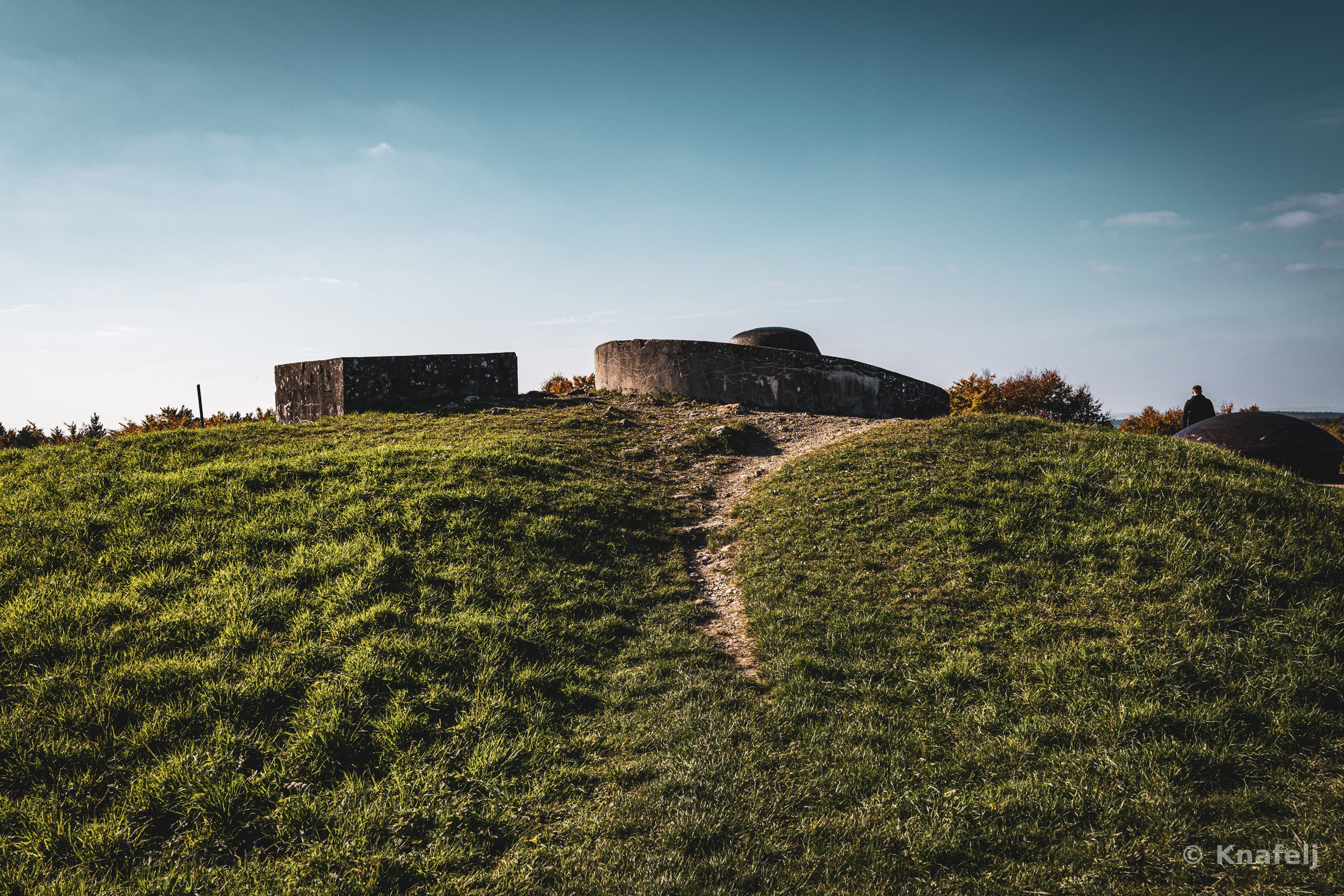 Old concrete military bunkers and rounded steel gun turrets on top of a grass-covered hill, with a worn dirt path leading up to them and a person standing at the right edge under a clear sky.