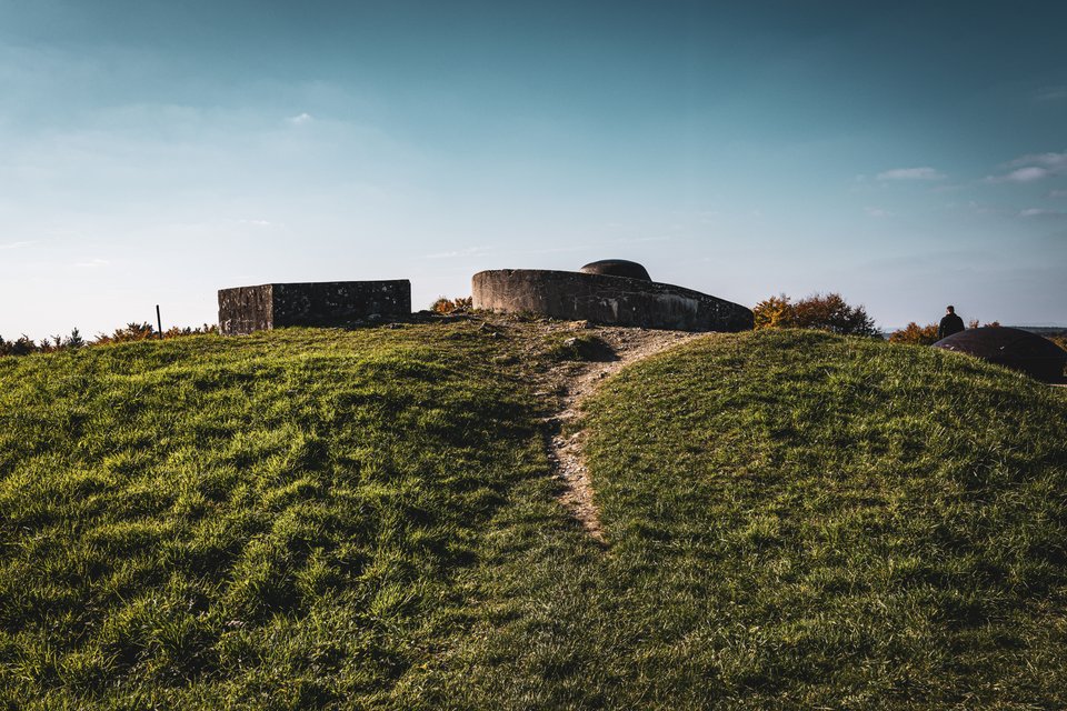 Old concrete military bunkers and rounded steel gun turrets on top of a grass-covered hill, with a worn dirt path leading up to them and a person standing at the right edge under a clear sky.