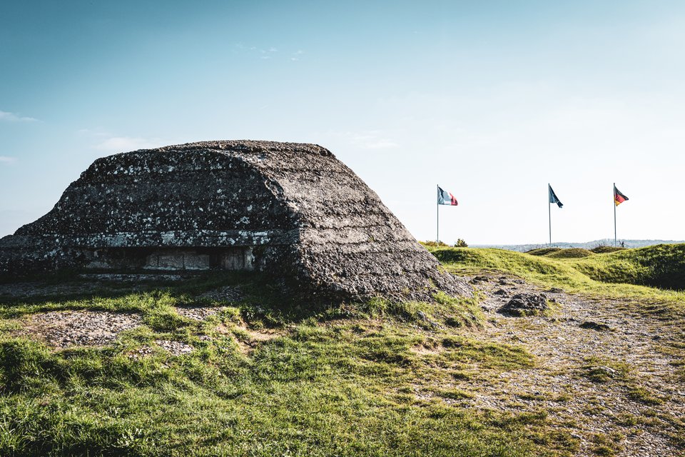 An old eroded concrete bunker on a grassy hilltop with three flags flying on poles in the background under a clear sky.