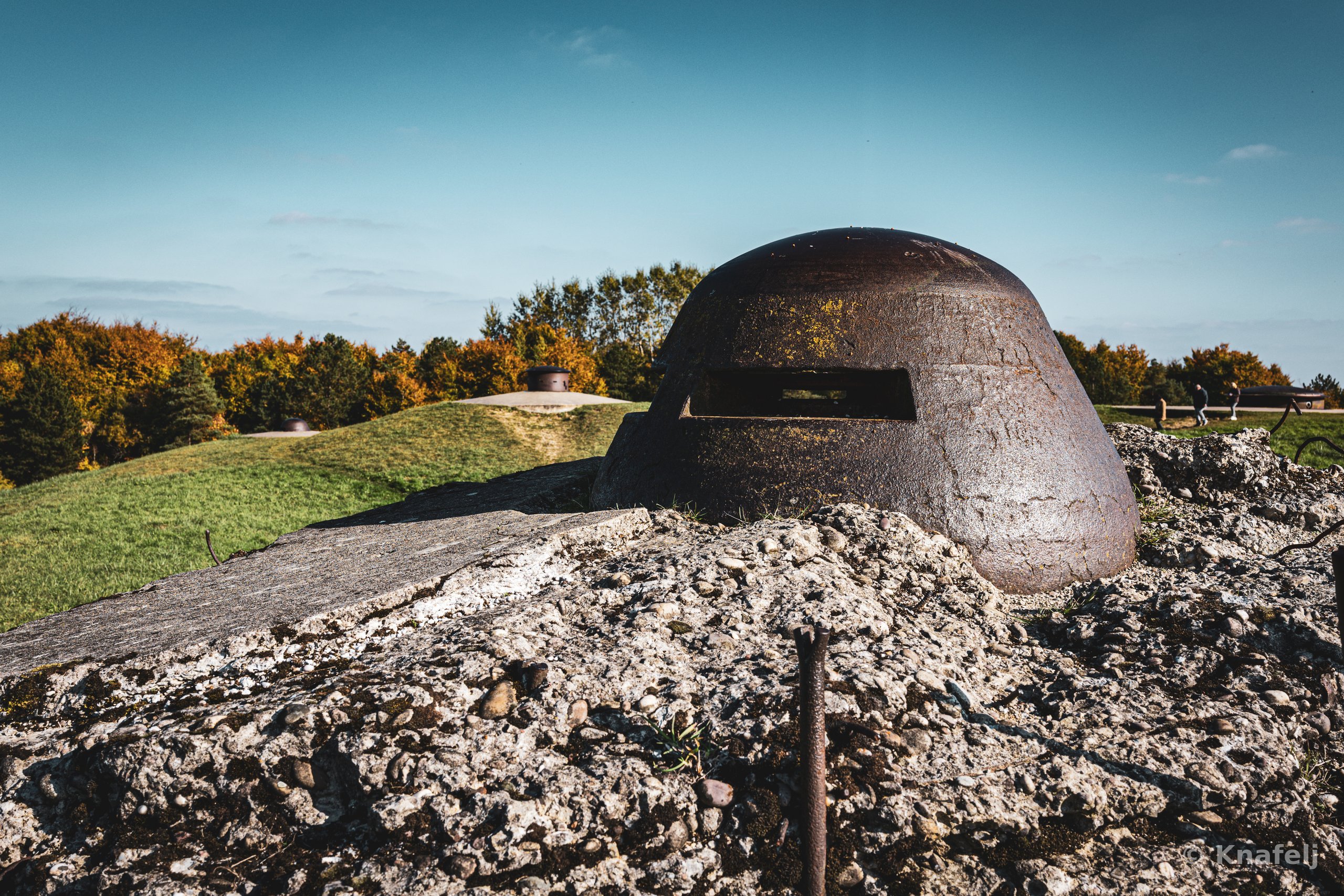 A rusted dome-shaped armored military cupola with a horizontal viewing slit, set on cracked concrete and gravel, surrounded by green grass and autumn-colored trees under a blue sky.
