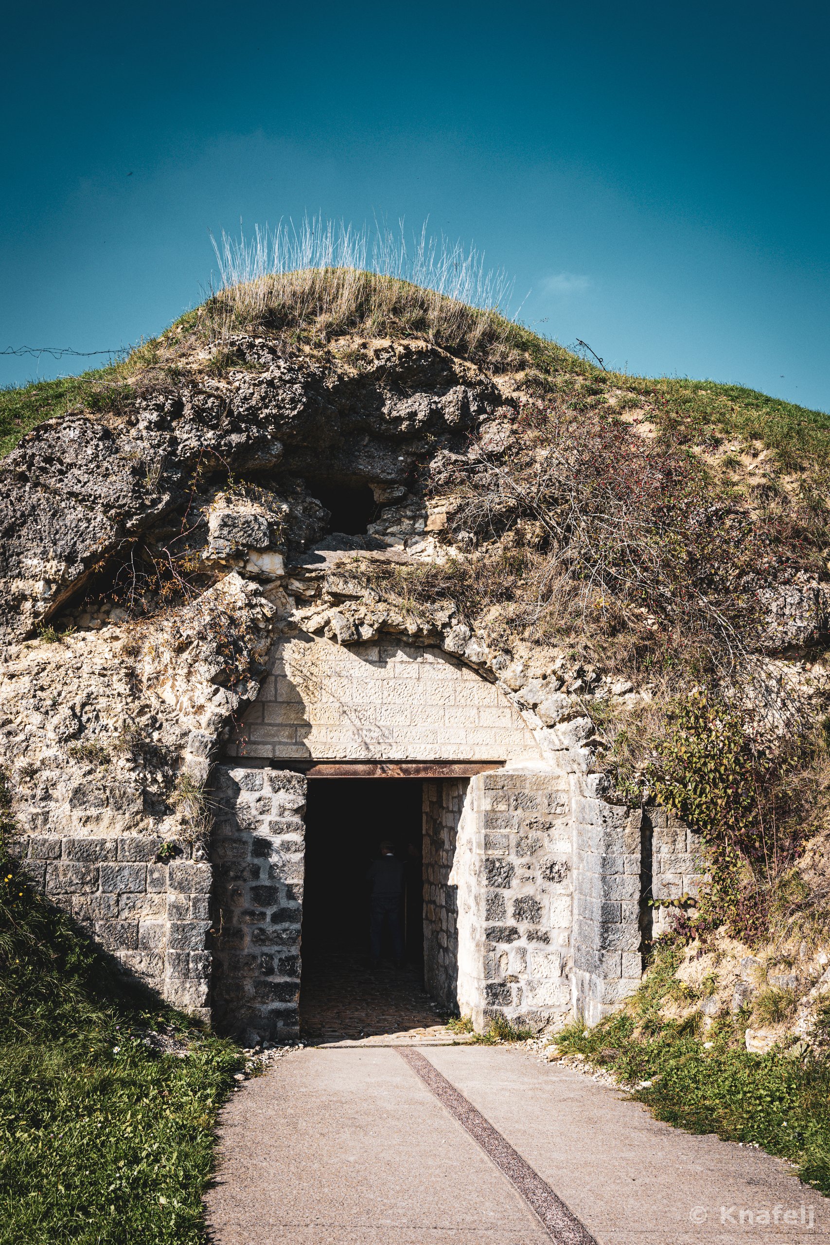 A stone entrance built into a grassy hill leads into a dark doorway, with crumbling rock above and a paved path approaching under a clear blue sky.