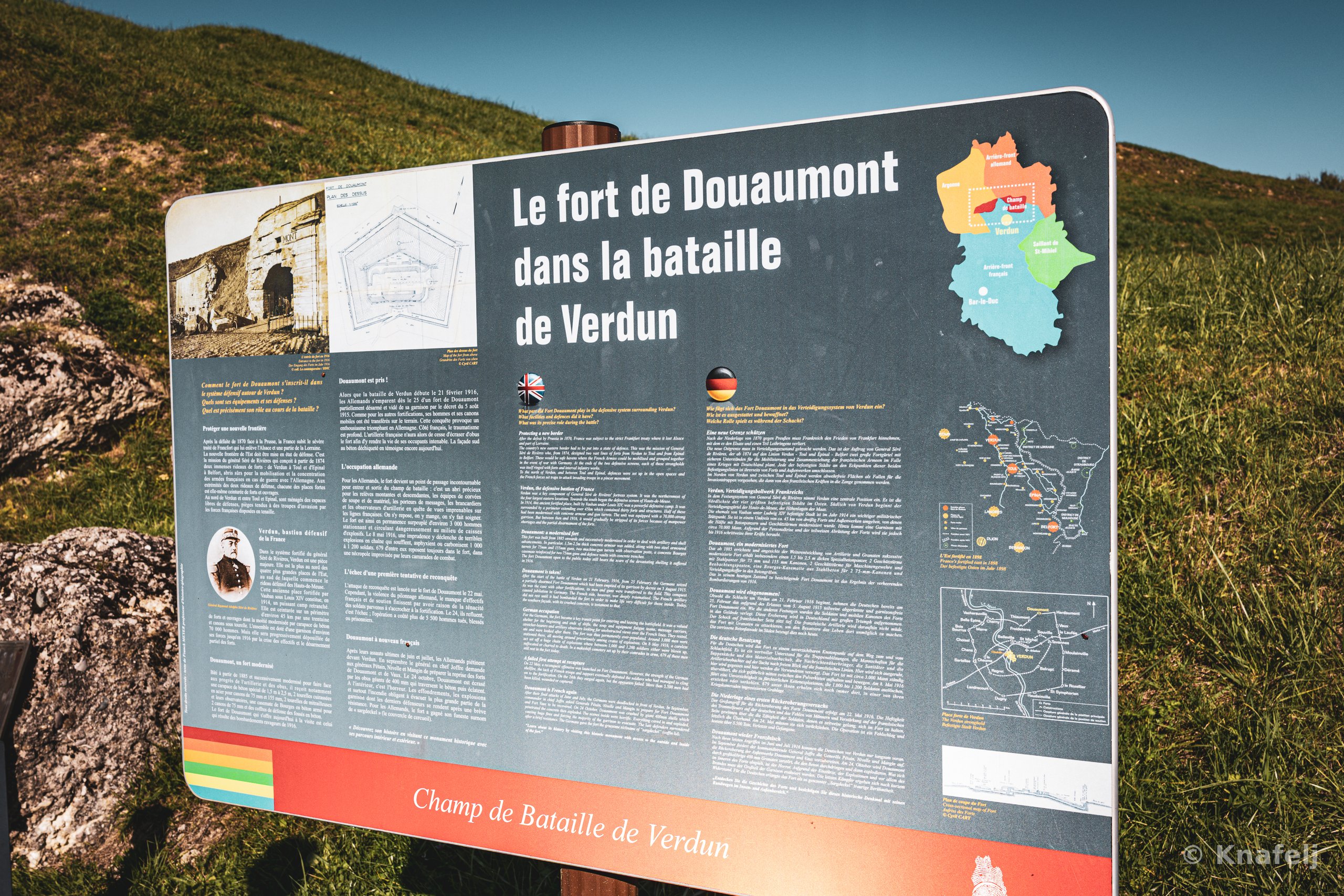 A large outdoor interpretive sign titled 'Le fort de Douaumont dans la bataille de Verdun' with maps, historical photos, and multilingual text, set against a green grassy hill under a blue sky.