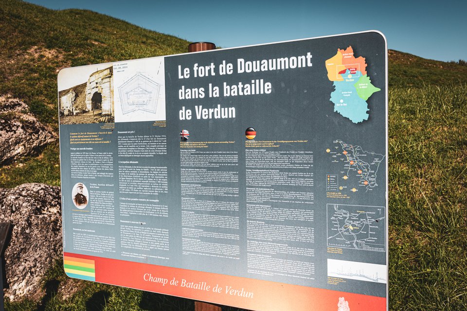 A large outdoor interpretive sign titled 'Le fort de Douaumont dans la bataille de Verdun' with maps, historical photos, and multilingual text, set against a green grassy hill under a blue sky.