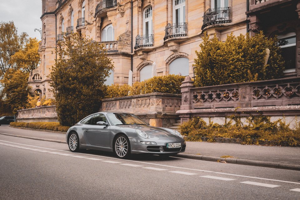 A silver Porsche sports car parked on the side of an empty road in front of a grand stone mansion with carved facades, balconies, and surrounding shrubbery in warm autumn tones.