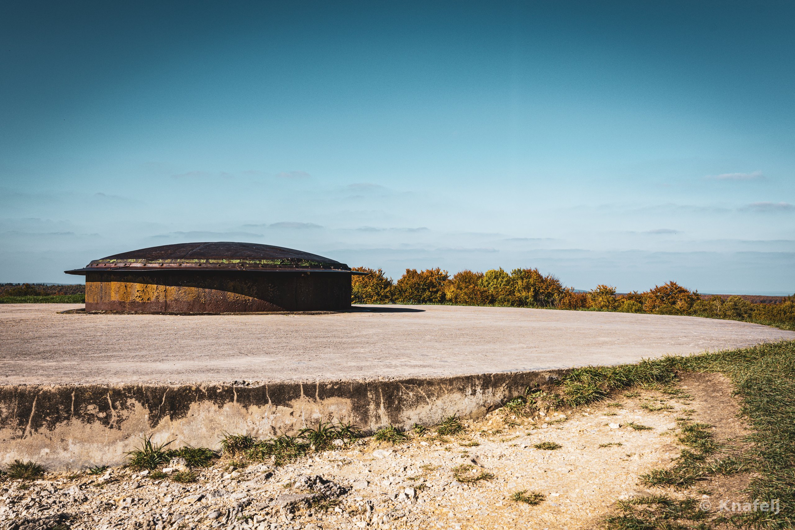 A rusted dome-shaped steel gun turret embedded in a circular concrete platform on a hilltop, with green and golden shrubs behind it and a clear blue sky above.