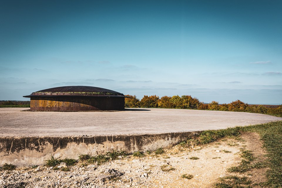 A rusted dome-shaped steel gun turret embedded in a circular concrete platform on a hilltop, with green and golden shrubs behind it and a clear blue sky above.