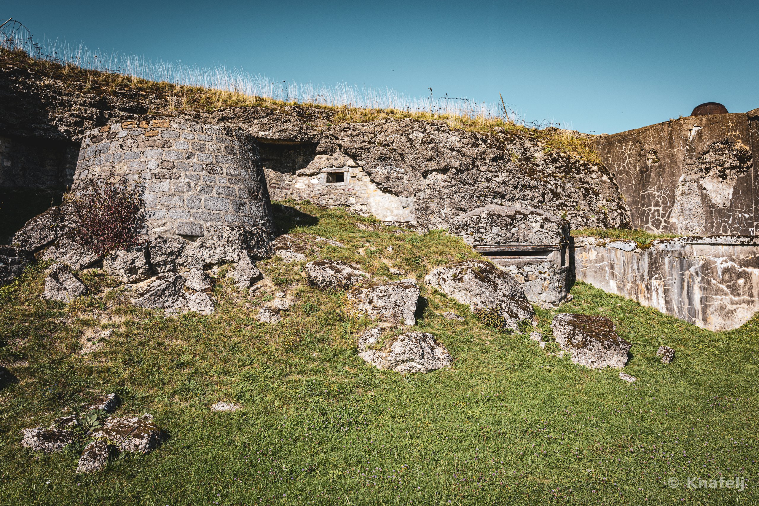 Old stone fort walls with grass growing on top, set against a deep blue sky, with rocks scattered across a green grassy hill in the foreground.