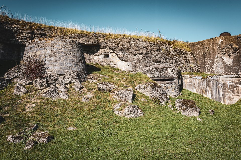 Old stone fort walls with grass growing on top, set against a deep blue sky, with rocks scattered across a green grassy hill in the foreground.
