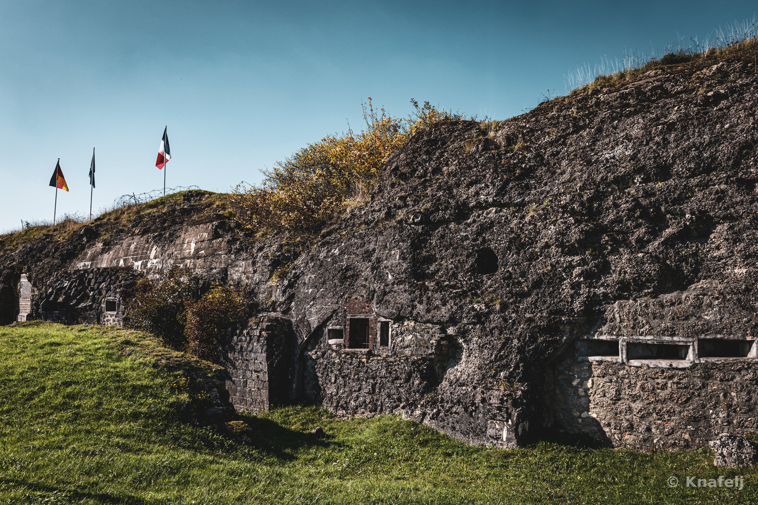 An old stone fortress embedded in a grassy hill with three flags flying on top, including a German and French flag, under a clear blue sky. The weathered ruins feature small windows and arched openings, with green grass in the foreground.
