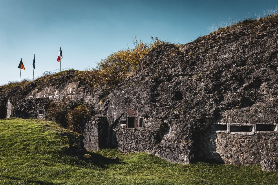 An old stone fortress embedded in a grassy hill with three flags flying on top, including a German and French flag, under a clear blue sky. The weathered ruins feature small windows and arched openings, with green grass in the foreground.
