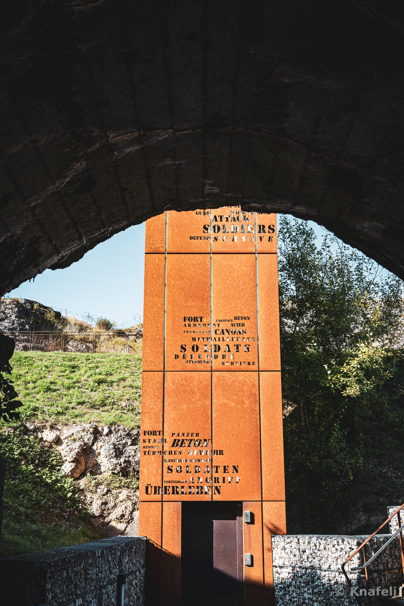 A rust-colored steel tower covered in stenciled text in English, French, and German is viewed through a large stone arch, with green grass, trees, and a clear blue sky in the background.