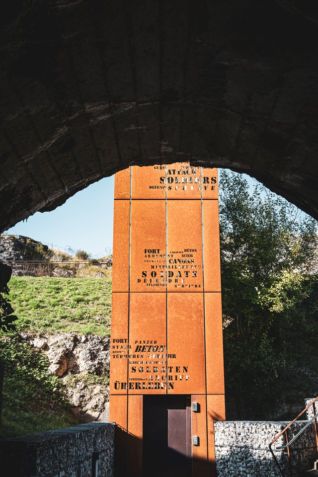 A rust-colored steel tower covered in stenciled text in English, French, and German is viewed through a large stone arch, with green grass, trees, and a clear blue sky in the background.