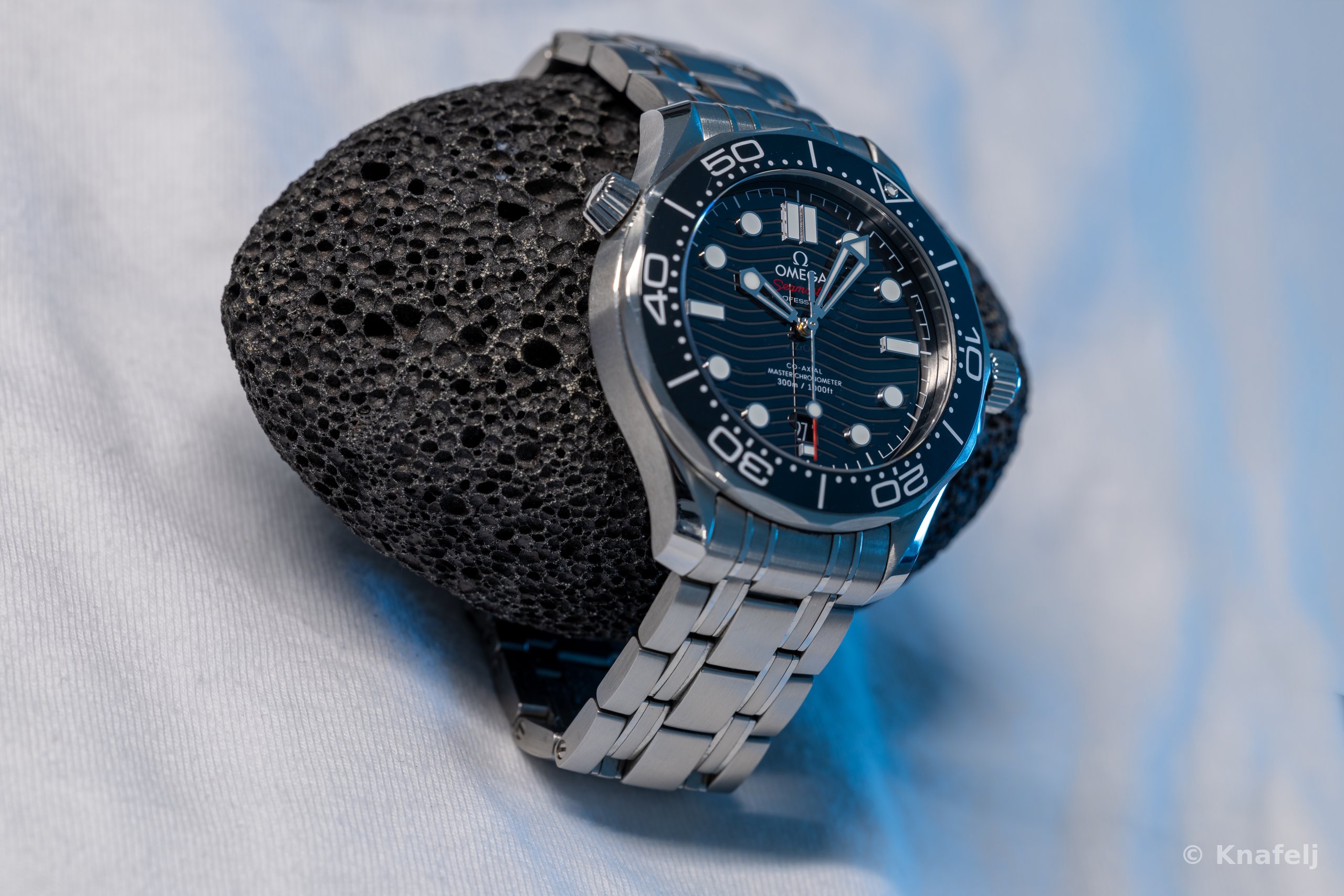 A luxury wristwatch with a blue wave-pattern dial, blue bezel, and stainless steel bracelet propped against a black porous lava rock on a light fabric surface.