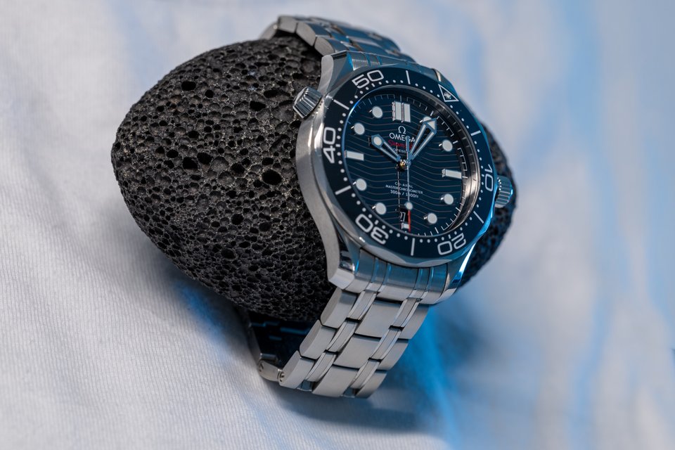 A luxury wristwatch with a blue wave-pattern dial, blue bezel, and stainless steel bracelet propped against a black porous lava rock on a light fabric surface.