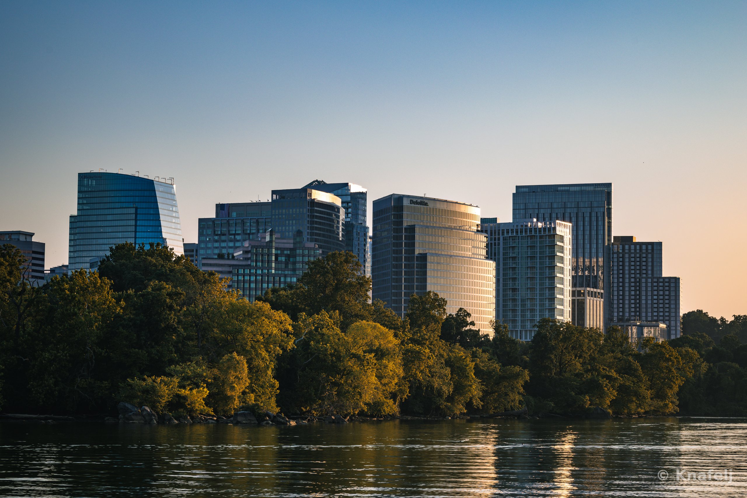 A city skyline of glass office towers illuminated by warm sunset light, set behind a dense line of green trees along a river, with the water reflecting the scene in the foreground.
