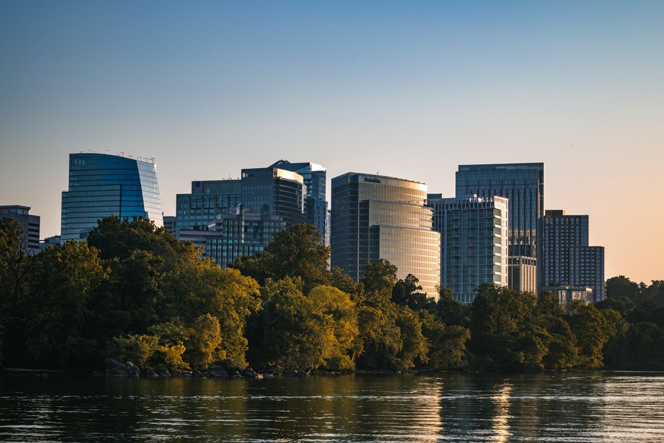 A city skyline of glass office towers illuminated by warm sunset light, set behind a dense line of green trees along a river, with the water reflecting the scene in the foreground.