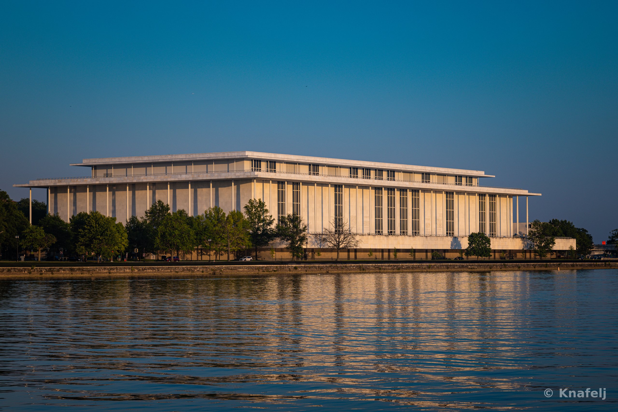 A large rectangular marble building with tall vertical columns sits along a river's edge, bathed in warm golden light under a clear blue sky, with trees at its base and its reflection in the rippling water below.