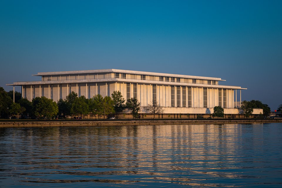 A large rectangular marble building with tall vertical columns sits along a river's edge, bathed in warm golden light under a clear blue sky, with trees at its base and its reflection in the rippling water below.