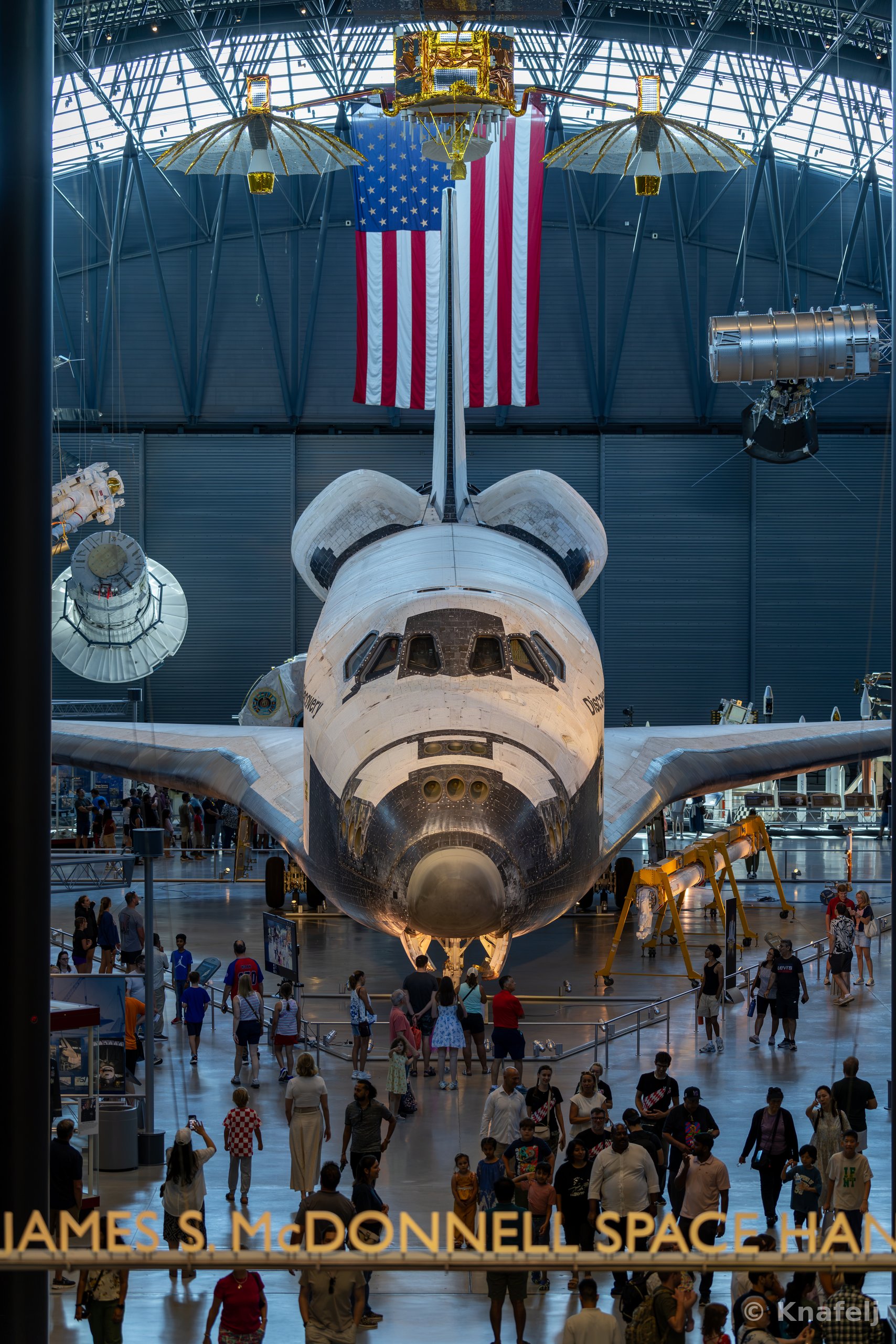 A retired NASA space shuttle displayed nose-forward in a museum hangar, with a large American flag hanging behind it and many visitors walking on the floor beneath the aircraft.