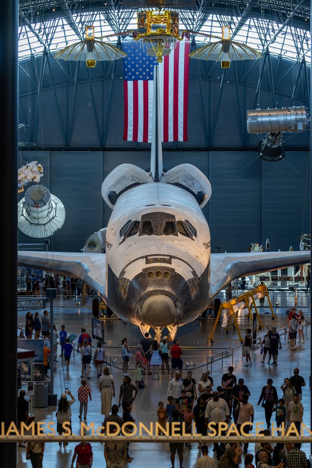 A retired NASA space shuttle displayed nose-forward in a museum hangar, with a large American flag hanging behind it and many visitors walking on the floor beneath the aircraft.
