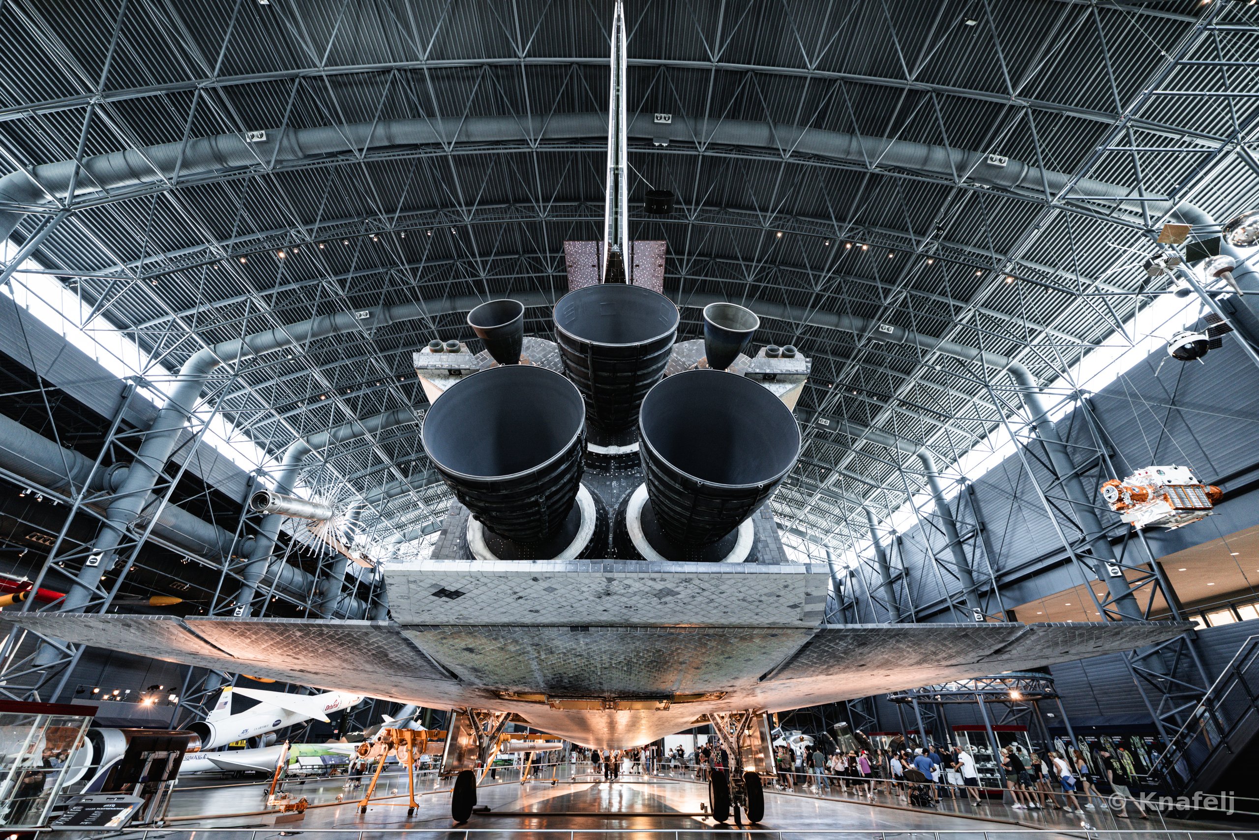 Rear view of a space shuttle's three main engines and heat-tiled underside, suspended in a large museum hangar with a curved steel lattice ceiling and crowds of people walking below.