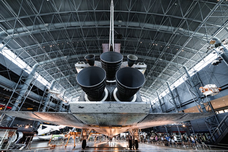 Rear view of a space shuttle's three main engines and heat-tiled underside, suspended in a large museum hangar with a curved steel lattice ceiling and crowds of people walking below.