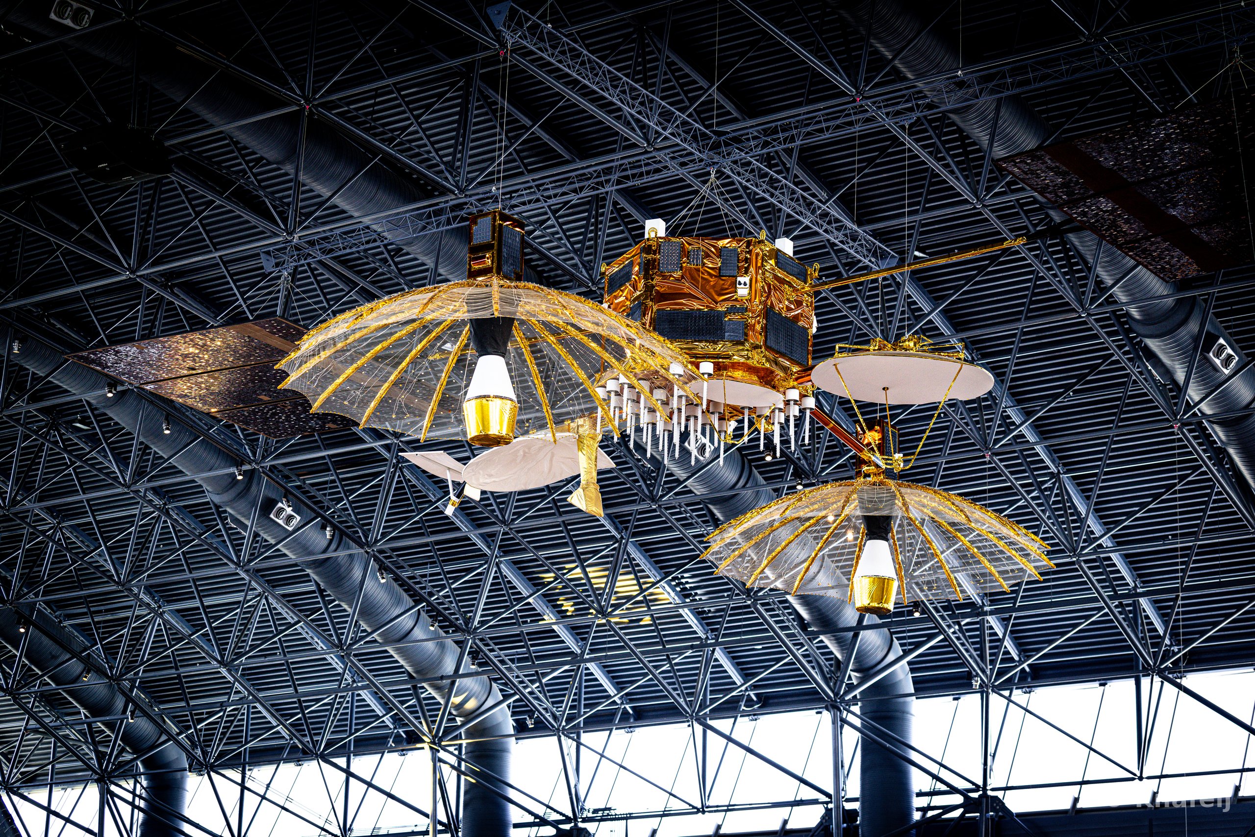 A gold and black spacecraft model with large parabolic mesh antennas and solar panels suspended from a dark geometric steel-framed ceiling indoors.