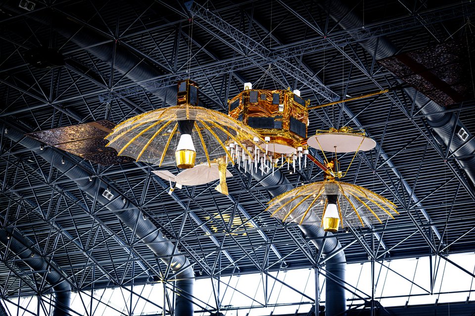 A gold and black spacecraft model with large parabolic mesh antennas and solar panels suspended from a dark geometric steel-framed ceiling indoors.