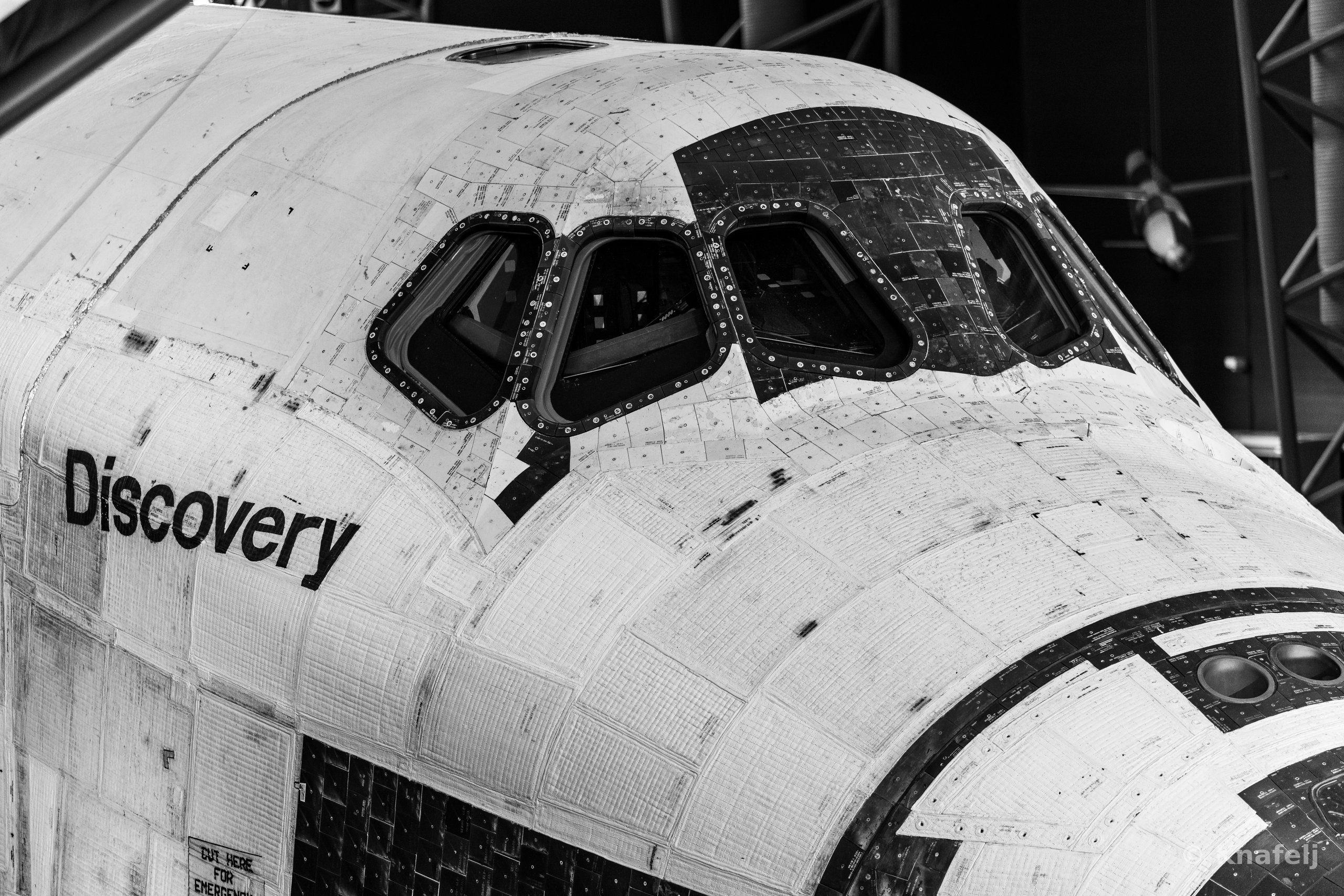 Black and white photograph of the forward section of Space Shuttle Discovery, showing its cockpit windows, textured heat-shield tiles, and the name 'Discovery' painted on the fuselage.