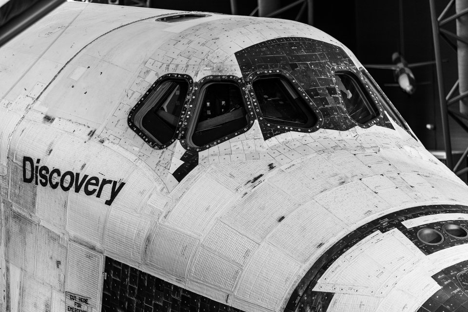 Black and white photograph of the forward section of Space Shuttle Discovery, showing its cockpit windows, textured heat-shield tiles, and the name 'Discovery' painted on the fuselage.