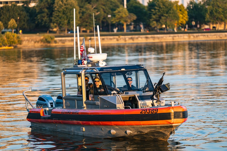 A U.S. Coast Guard patrol boat numbered 29389 with several crew members aboard, equipped with a mounted machine gun, radar dome, and American flag, moving across calm water with trees reflected in the background.