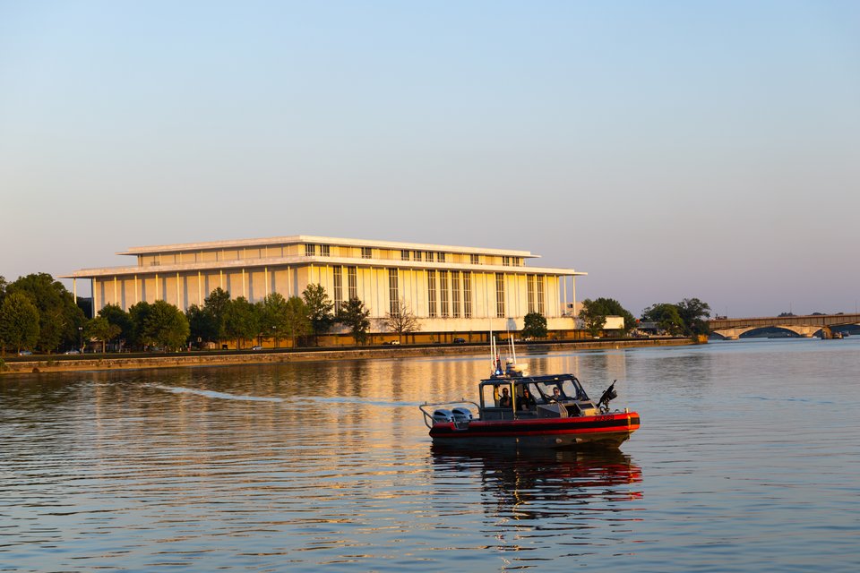 A red and black boat with people aboard travels on a calm river at sunset, with a large white modern building and a bridge visible along the far shoreline.