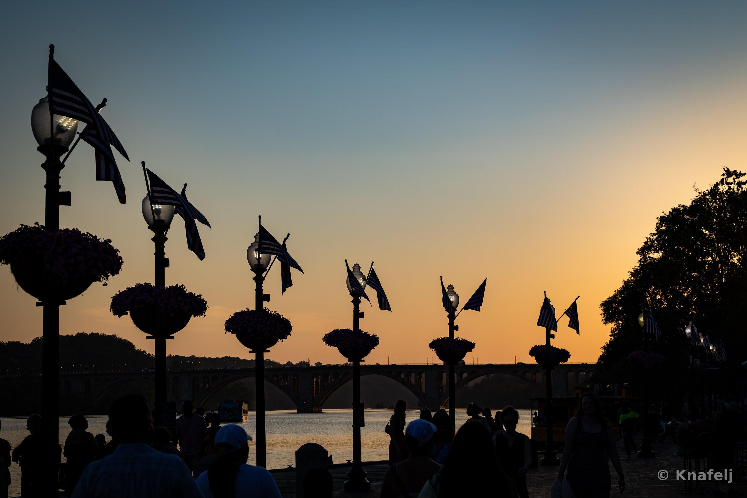 Silhouettes of ornate lampposts with hanging flags and flower baskets along a river walkway at sunset, with an arched stone bridge and people in the foreground against an orange and blue sky.