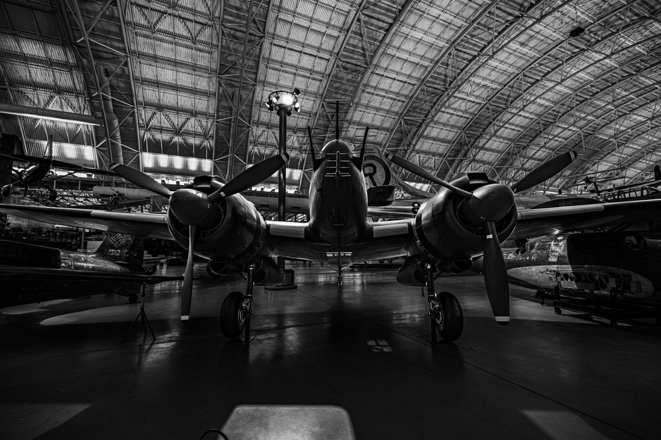 Black and white photo of a twin-engine propeller aircraft displayed inside a large museum hangar with a curved lattice ceiling and other planes in the background.