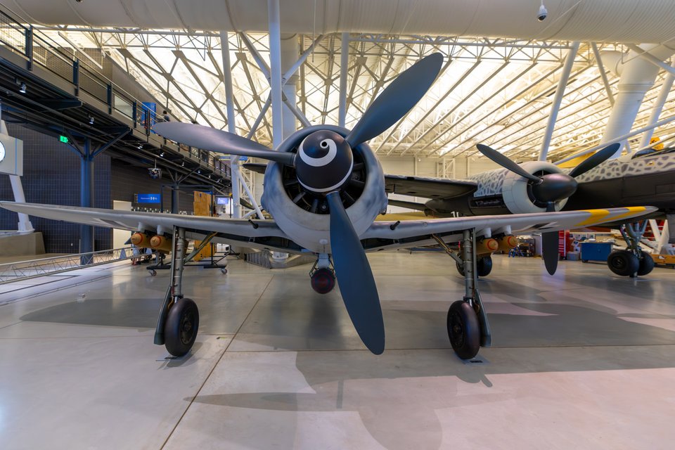 Front view of a gray World War II Focke-Wulf 190 fighter plane with a black spinner and four-bladed propeller, parked indoors in a large aviation museum with other aircraft and a lattice ceiling behind it.
