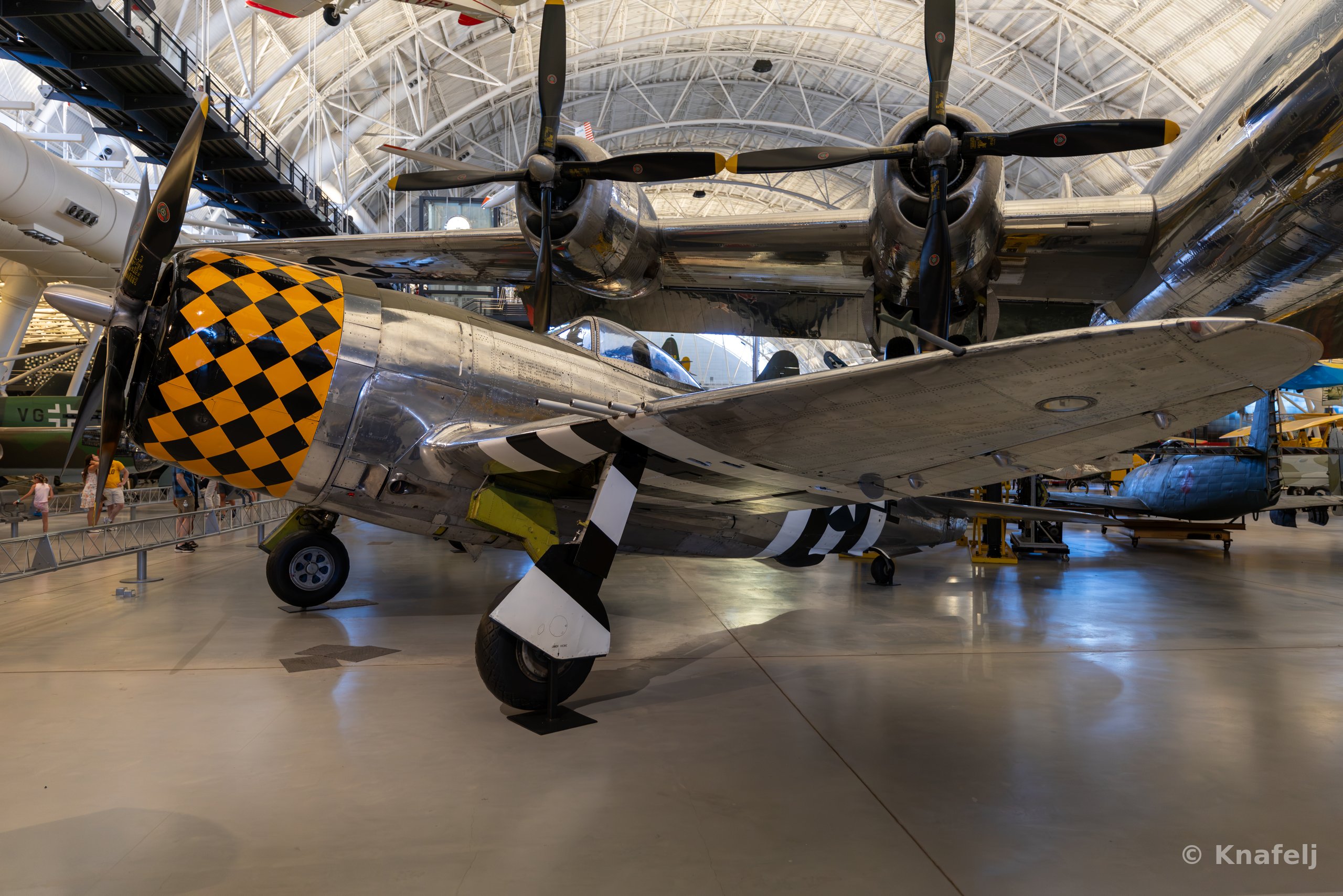 A silver World War II fighter aircraft with a yellow and black checkerboard engine cowling and black-and-white striped propeller blades parked indoors in a large museum hangar, with other aircraft and an arched ceiling visible in the background.