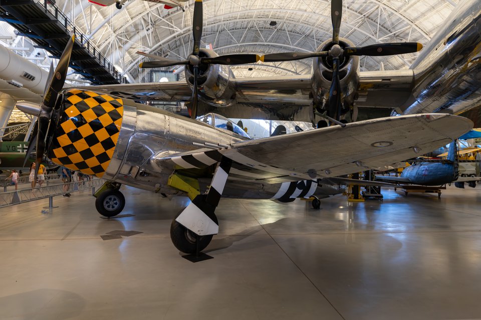 A silver World War II fighter aircraft with a yellow and black checkerboard engine cowling and black-and-white striped propeller blades parked indoors in a large museum hangar, with other aircraft and an arched ceiling visible in the background.