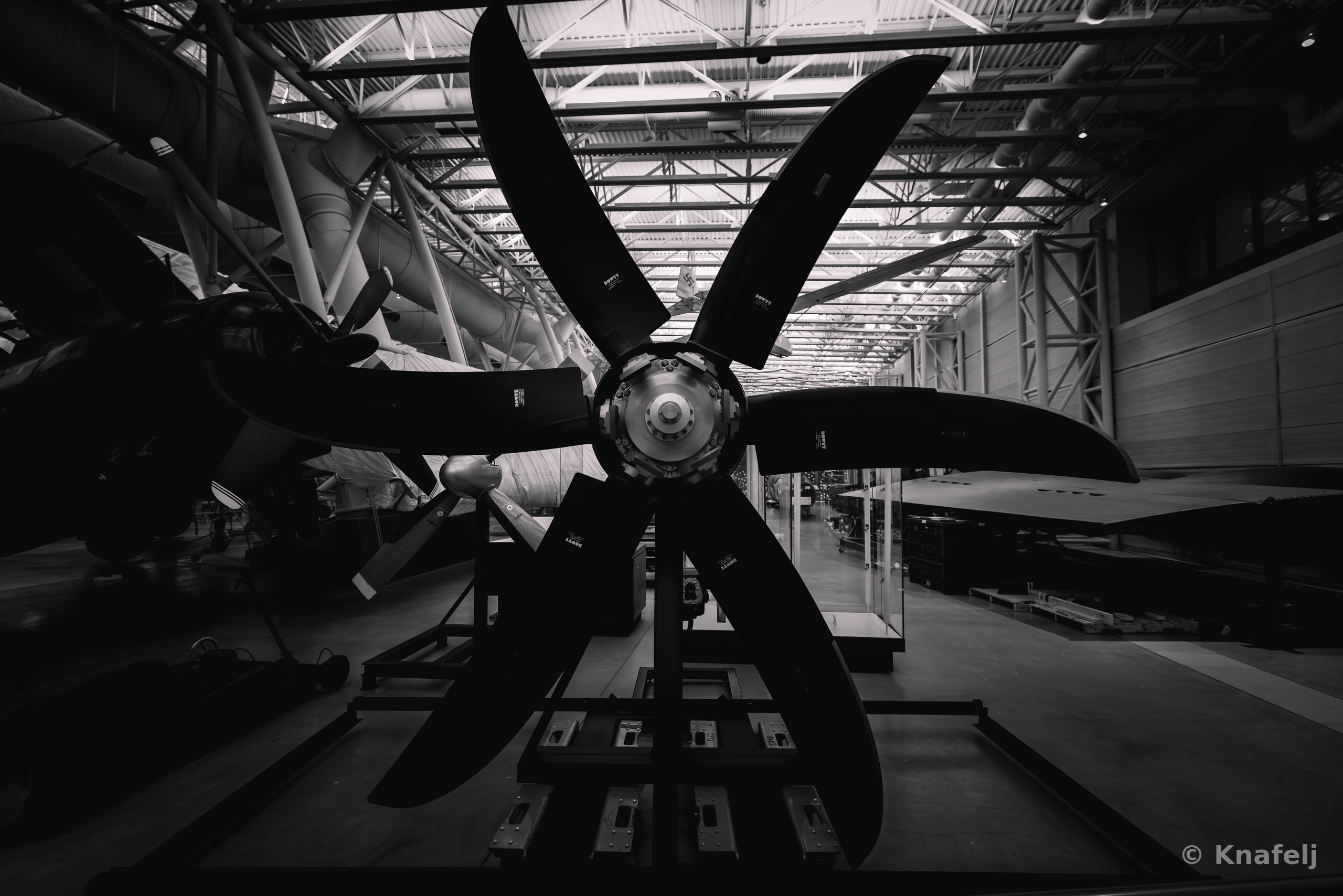 Black and white image of a large six-bladed aircraft propeller mounted on a stand inside a museum hangar, with steel roof trusses and aircraft visible in the background.