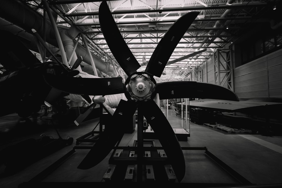 Black and white image of a large six-bladed aircraft propeller mounted on a stand inside a museum hangar, with steel roof trusses and aircraft visible in the background.