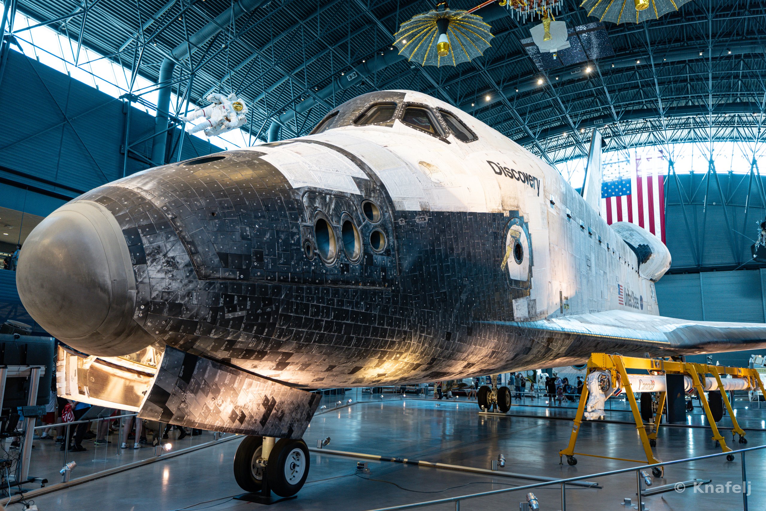 Space Shuttle Discovery viewed from the front left, showing its scorched black and white heat-shield tiles, cockpit windows, and landing gear, displayed indoors in a large museum hangar with an American flag and hanging satellites behind it.