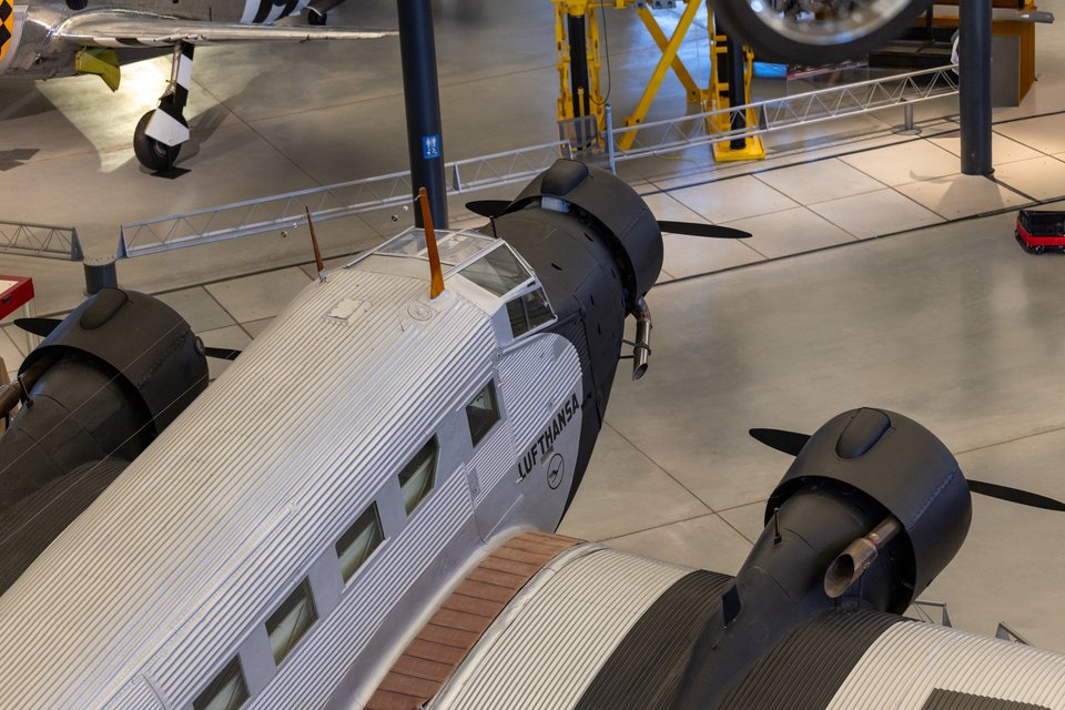 Overhead view of a silver Lufthansa Junkers Ju 52 aircraft with corrugated metal skin, showing the cockpit, three dark engines with propellers, and rows of cabin windows, parked on a concrete floor in a museum among other planes.