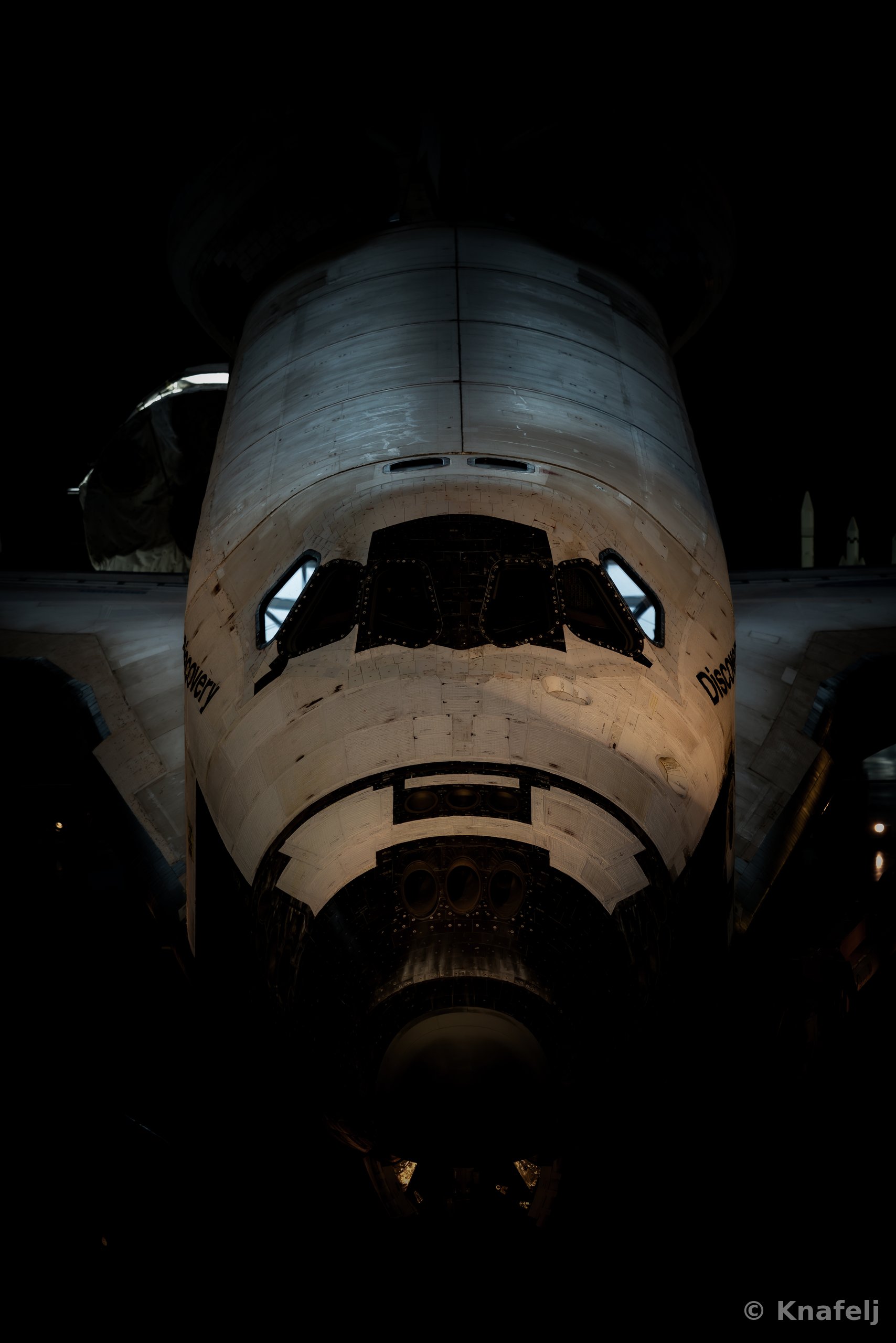 Front view of the Space Shuttle Discovery's nose and cockpit windows, illuminated against a dark background, showing weathered thermal tiles and the name Discovery on the fuselage.