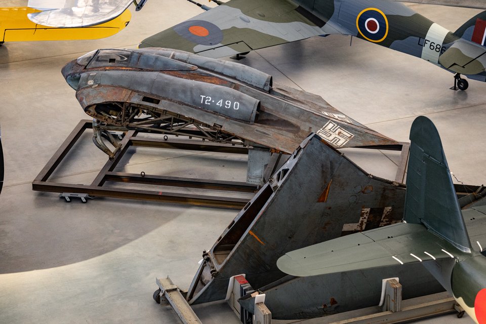 A weathered, incomplete Horten Ho 229 flying wing fuselage marked T2-490 sitting on a steel frame dolly on a concrete museum floor, with parts of other aircraft nearby.