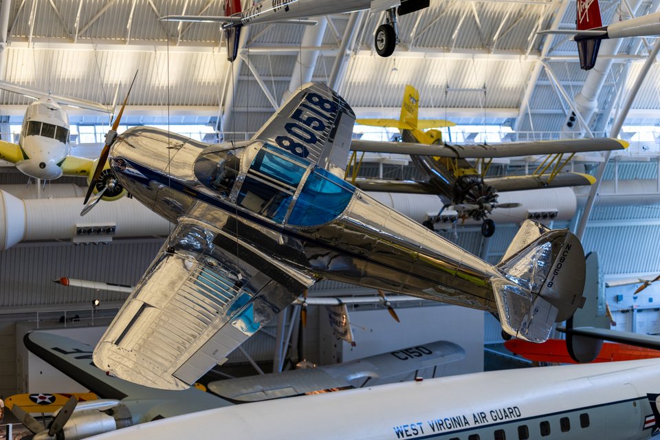 A shiny silver metal airplane with number 80518 painted on its tail and blue cockpit, suspended from the ceiling of an aircraft museum, with other planes and a West Virginia Air Guard aircraft visible in the background.